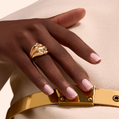 A hand with Manucurist Milky Pink gel polish nails wears a gold ring with three diamonds, resting on a white fabric outfit with a shiny gold belt.