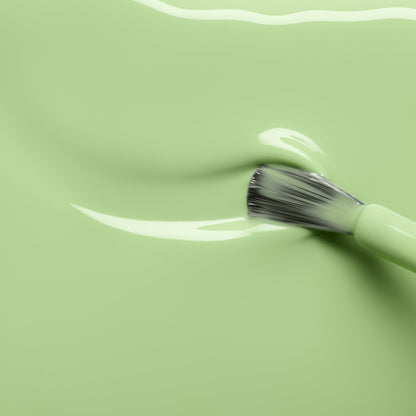 A makeup brush glides through the smooth, glossy Milky Matcha cream by Manucurist, its light green hue reminiscent of matcha nail polish, creating a gentle wave across the surface.