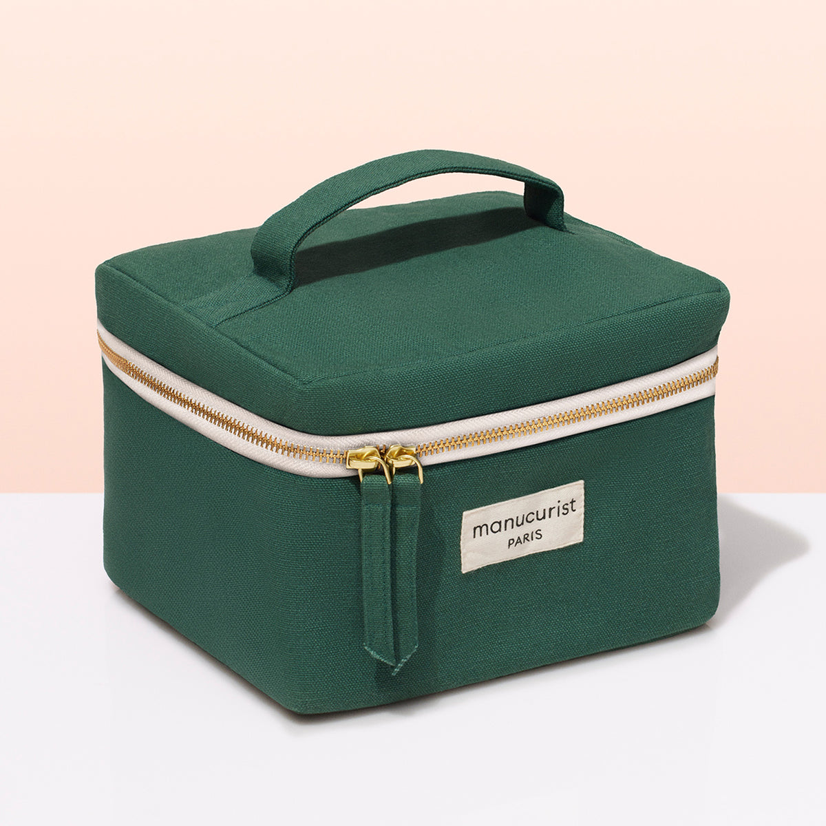 The Manucurist Vanity Pouch is a green fabric case with a handle, gold zipper, and white Manucurist PARIS label—ideal for storing nail polish—shown on a white and light peach background.