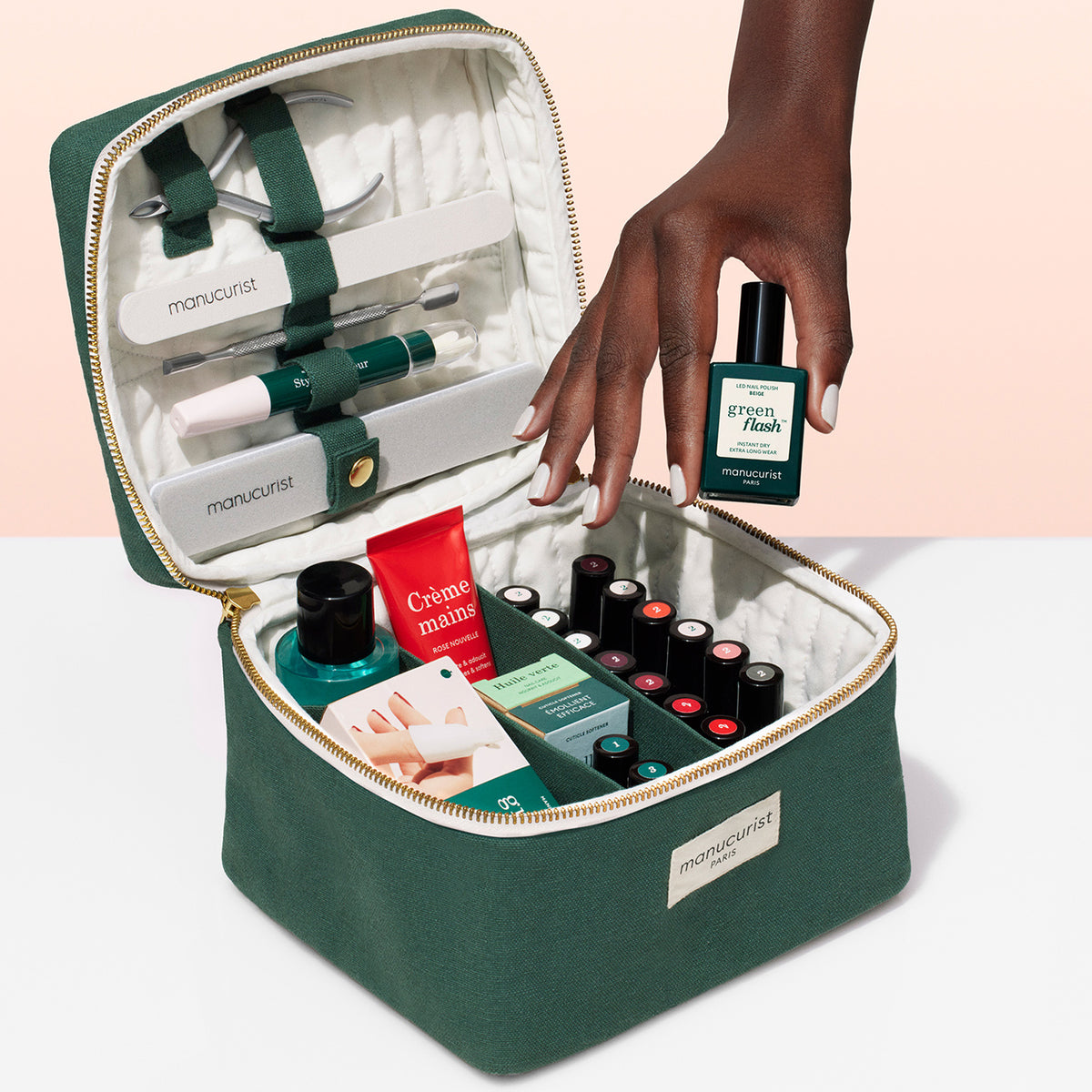 A hand holds a bottle of green nail polish above an open Manucurist Vanity Pouch, displaying neatly organized bottles, hand cream, nail files, and other manicure tools inside this stylish zippered kit.