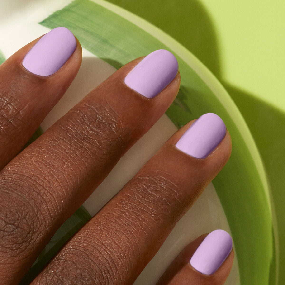 A close-up of a dark-skinned hand with neatly manicured nails painted in Manucurist’s Lisa Lilas gel polish, resting on a round object with green and white patterns against a light green background.