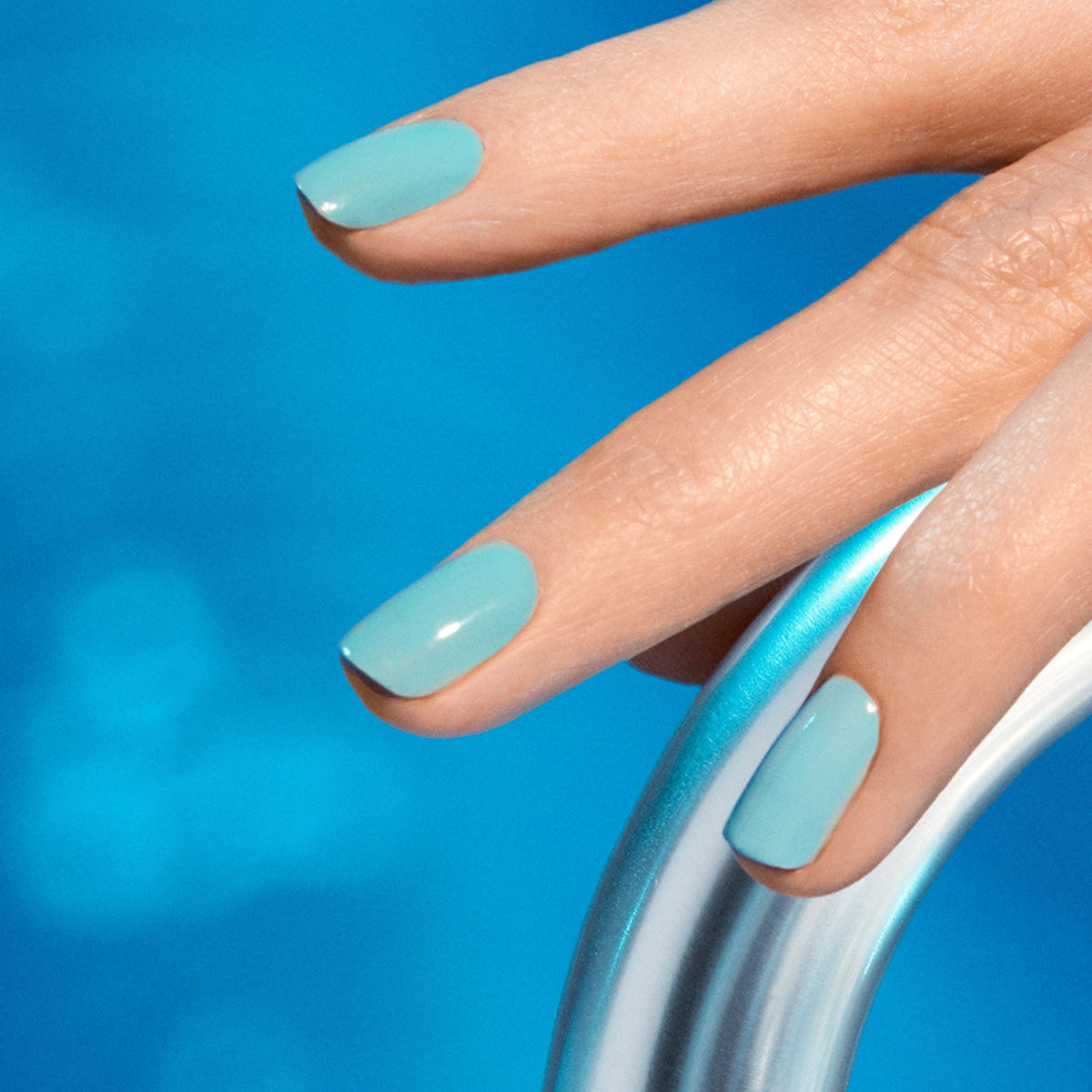 A close-up of a hand shows nails painted with Riviera gel polish by Manucurist, resting on a metallic pool ladder against clear blue water in the background.