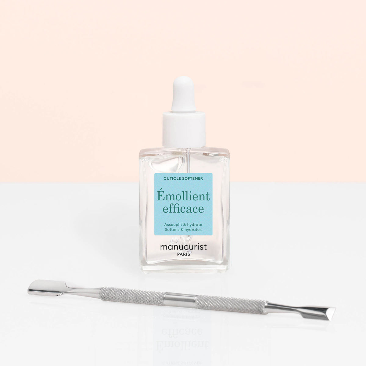 A bottle of Manucurist Hydrating Cuticle Remover with a dropper cap stands behind a double-ended metal cuticle pusher, ideal for manicures, on a white surface and light pink background.