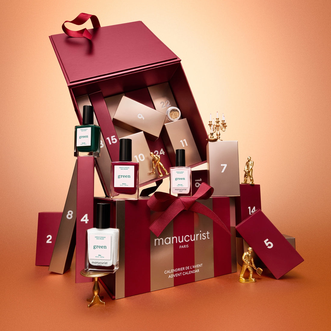 A burgundy “Advent Calendar Time to Shine!” box by Manucurist, featuring numbered drawers filled with manicured nail polishes, nail care essentials, and small gold figurines, displayed on a peach background.