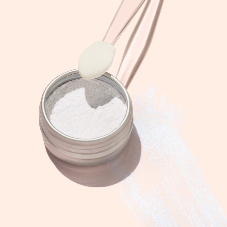 An open container of Manucurist Blue Glazed Powder rests on a pale surface, with a clear spoon above it holding the product. Pearly blue shimmer is scattered beside the container, reflecting light beautifully.