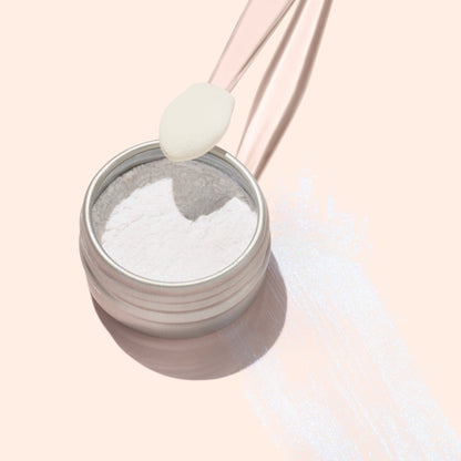 An open container of Manucurist Blue Glazed Powder rests on a pale surface, with a clear spoon above it holding the product. Pearly blue shimmer is scattered beside the container, reflecting light beautifully.