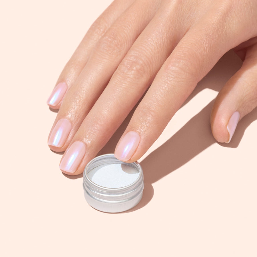 A hand with neatly manicured, iridescent pale pink nails—created using Manucurist Blue Glazed Powder—rests beside an open small round container of the powder on a light beige background.