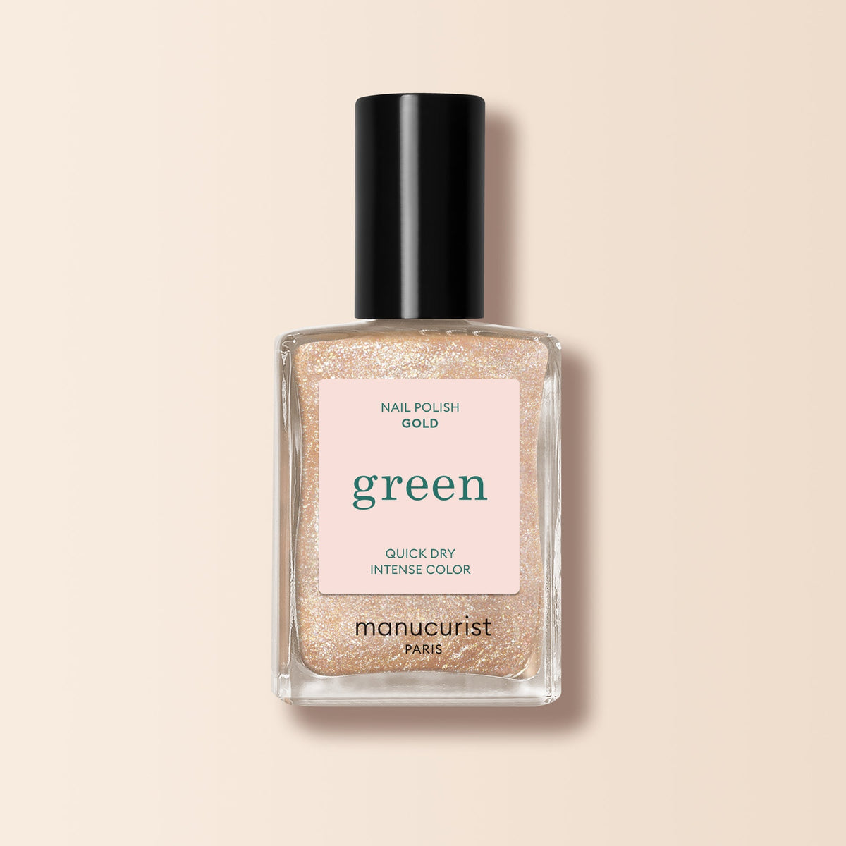A clear glass bottle of Manucurist Gold glitter nail polish with a black cap is shown on a soft beige background. The label reads Nail Polish Gold, green, Quick Dry Intense Color, and manucurist Paris.
