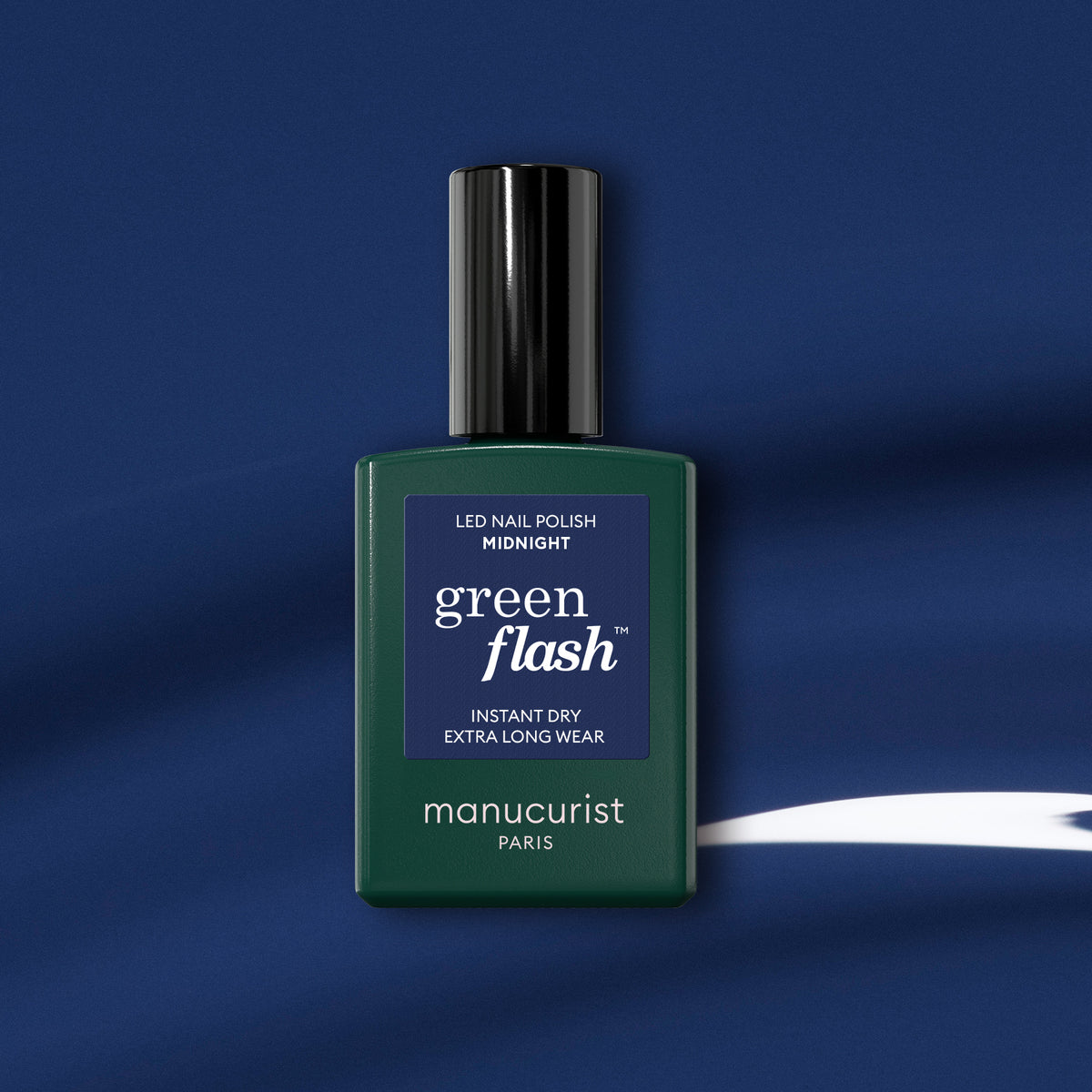 A Manucurist Midnight gel polish bottle stands against a blue background with a white streak, its dark green body, black cap, and blue label reflecting the rich midnight hue of the polish inside.