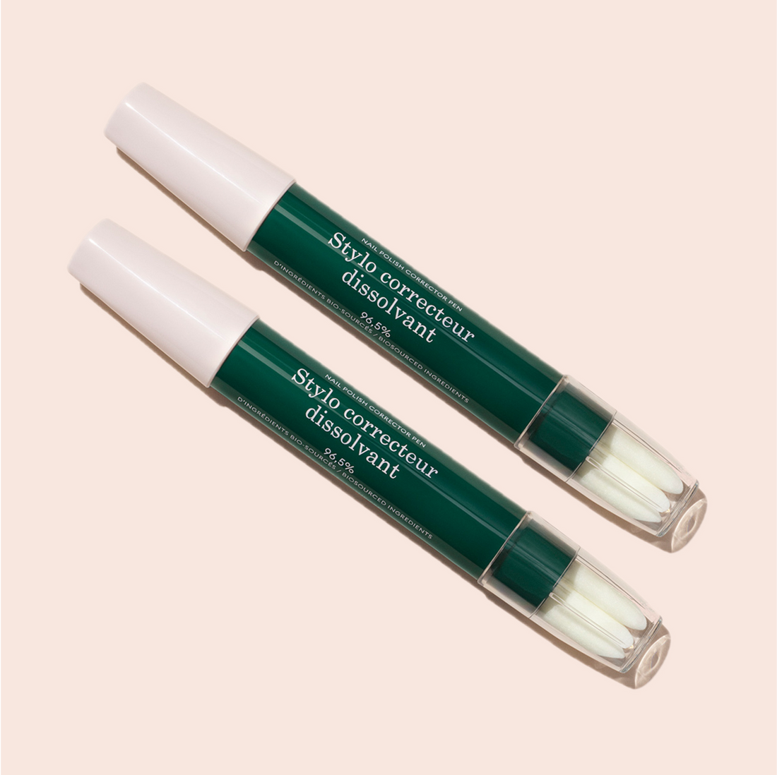Two green and white Nail Polish Corrector Pens labeled “Stylo correcteur dissolvant” lie diagonally on a light beige background. The clear caps reveal fine tips, ideal for cleaning up gel polish.