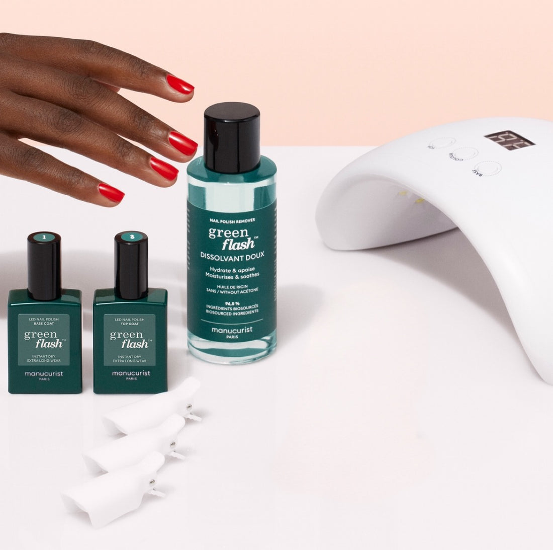 A hand with red-painted nails reaches for the Manucurist Welcome Kit Green Flash™ next to two green polishes, white nail clips, and a white LED lamp on a light pink surface.
