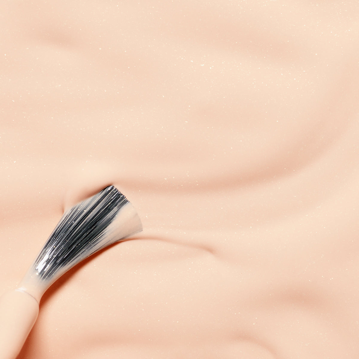 A makeup brush with white bristles rests on the smooth, peach-colored, slightly shimmery surface of Manucurist’s Joy the Bear Cub kids nail polish, creating gentle indents.