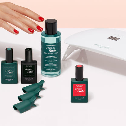 A hand with red nails is near the Manucurist Discovery Kit, featuring bottles of Green Flash nail products, three green nail clips, and a white LED lamp on a light surface—everything you need for an ultra-glossy manicure at home.