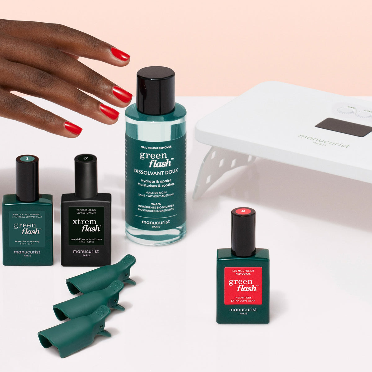 A hand with red nails reaches for Manucurist’s Discovery Kit—which includes green and red long-lasting nail polishes, a polish remover, green clips, and a white LED lamp—set against a pink and white background; it’s everything you need for an ultra-glossy manicure.