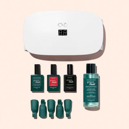 The Manucurist Discovery Kit, featuring a white nail lamp, three bottles of long-lasting nail polish, nail polish remover, and four green nail clips, is neatly arranged on a light pink background.