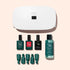 The Manucurist Discovery Kit, featuring a white nail lamp, three bottles of long-lasting nail polish, nail polish remover, and four green nail clips, is neatly arranged on a light pink background.