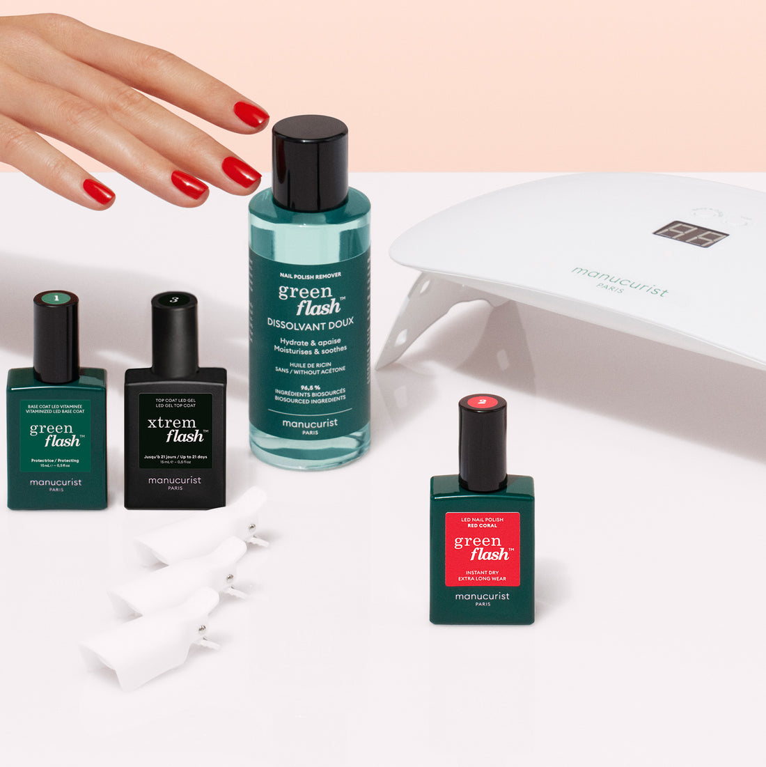 A hand with red nail polish is near the Manucurist Discovery Kit, which includes Green Flash, a vitaminized base coat, Xtreme nail products, a nail polish remover, a white LED nail lamp, and four white fingernail clips on a white surface.