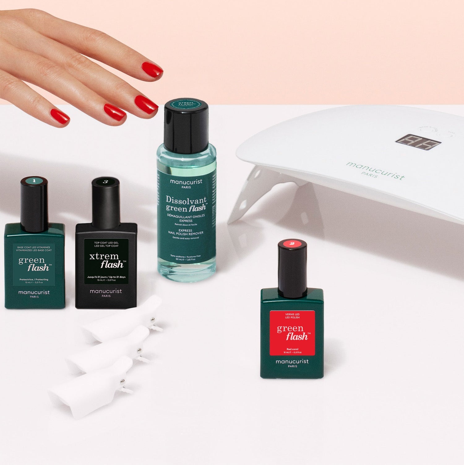 A hand with red polished nails reaches for Manucurist’s Discovery Kit, featuring red and green gel polishes, a vitaminized base coat, nail remover, white clips, and an LED lamp on a beige surface for an ultra-glossy manicure.