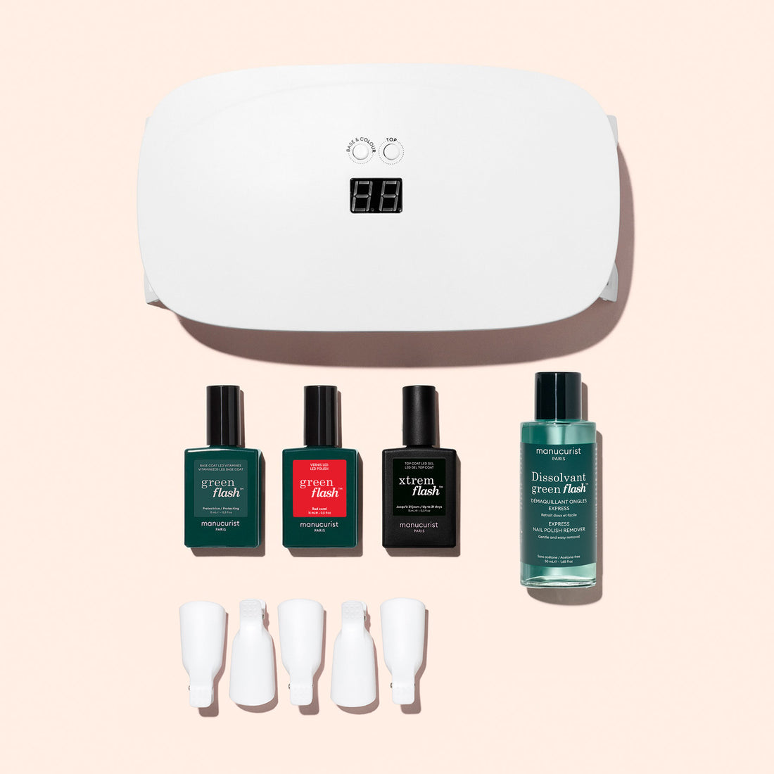 The Manucurist Discovery Kit, featuring a white LED nail lamp, three gel polishes, nail polish remover, and four clips, is arranged on a pale pink background for an ultra-glossy manicure.