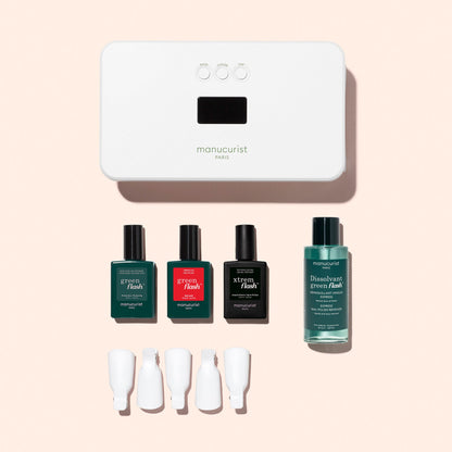 The Manucurist Discovery Kit includes a white LED nail lamp, three polishes (with vitaminized base coat), remover, and four white caps—everything you need for an ultra-glossy manicure, arranged on a chic pink background.