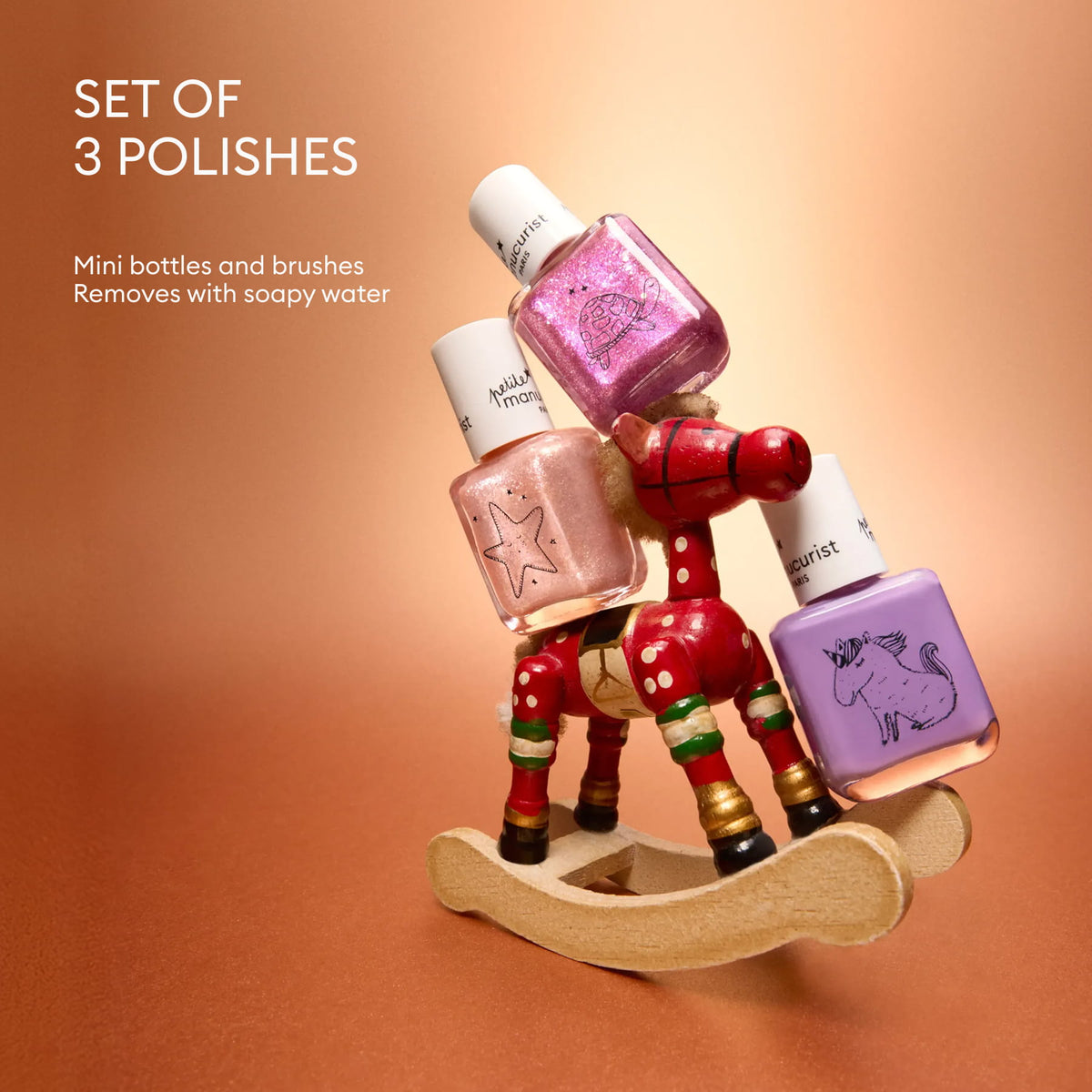 Three mini children’s nail polish bottles from Manucurist, in the Petite Manucurist Trio set, with shades of pink, purple, and peach, are balanced on a small wooden rocking horse. The background is a metallic rose gold. Text reads: Petite Manucurist Trio—Set of 3 polishes from Manucurist. Mini bottles and brushes made with natural origin ingredients. Removes easily with soapy water—perfect for holiday gifting.