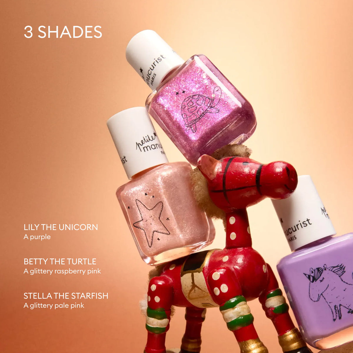 Introducing the Petite Manucurist Trio by Manucurist: three children’s nail polish bottles, crafted with natural origin ingredients, in playful shades—purple (LILY THE UNICORN), glittery raspberry pink (BETTY THE TURTLE), and glittery pale pink (STELLA THE STARFISH). Stacked atop a red wooden horse against a peach background, this set makes a perfect holiday gift for little ones who love fun colors!.