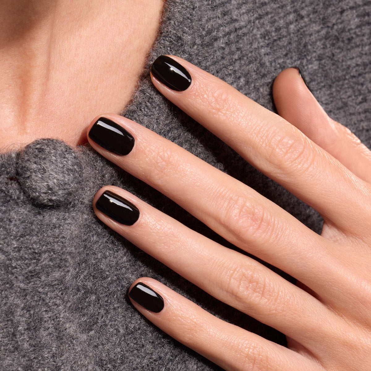 A hand with neatly manicured nails painted in Manucurist’s Licorice glossy black polish rests on a textured gray wool garment with a round button.