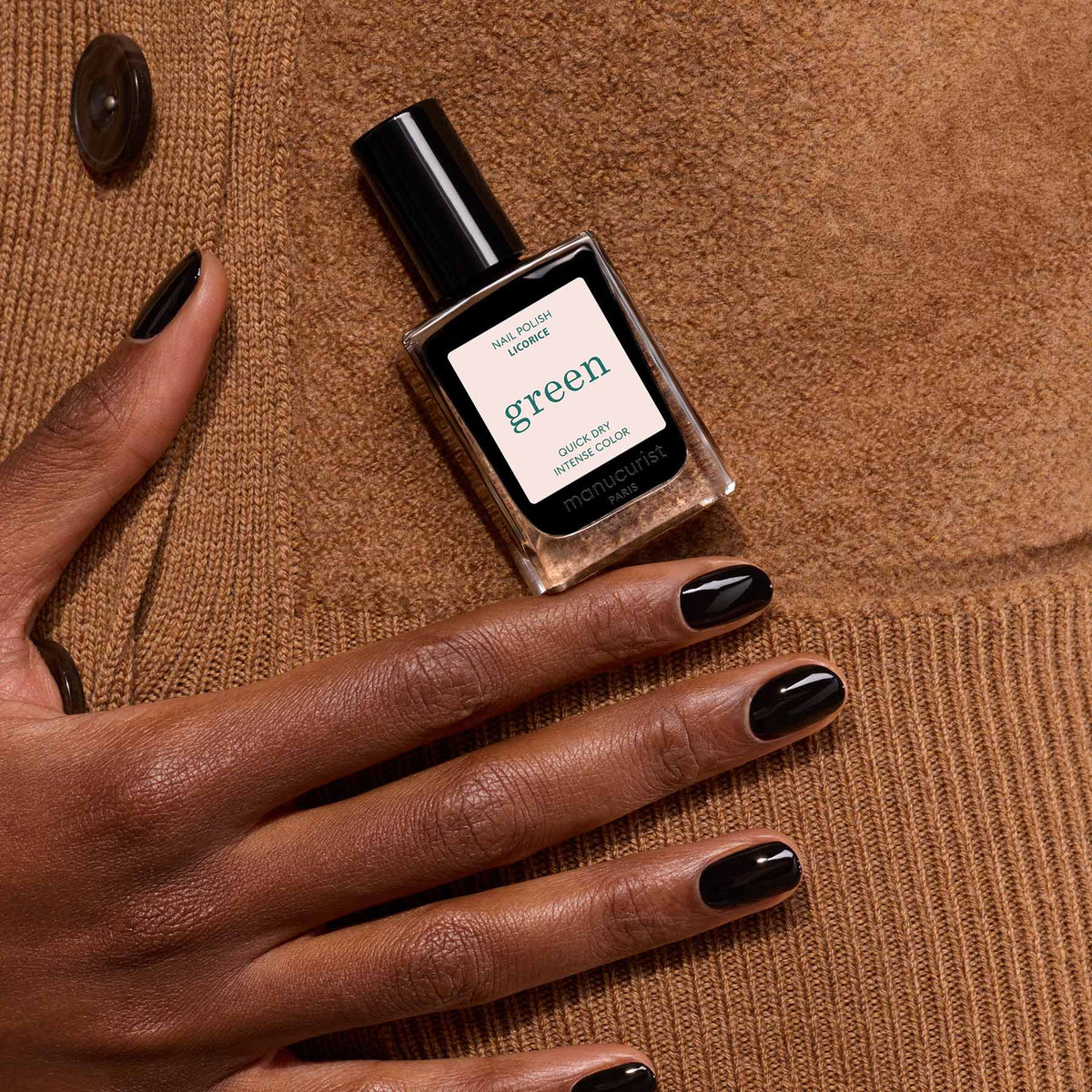 A hand with neatly manicured black nails holds a Manucurist Licorice bottle from the Green range, set against a brown suede jacket with button details.