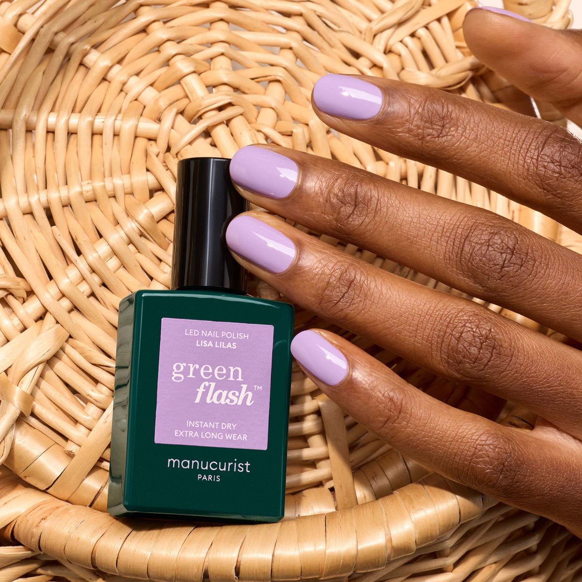 A hand with light purple nails rests on a woven surface beside a dark green Manucurist bottle, showcasing Lisa Lilas gel polish.