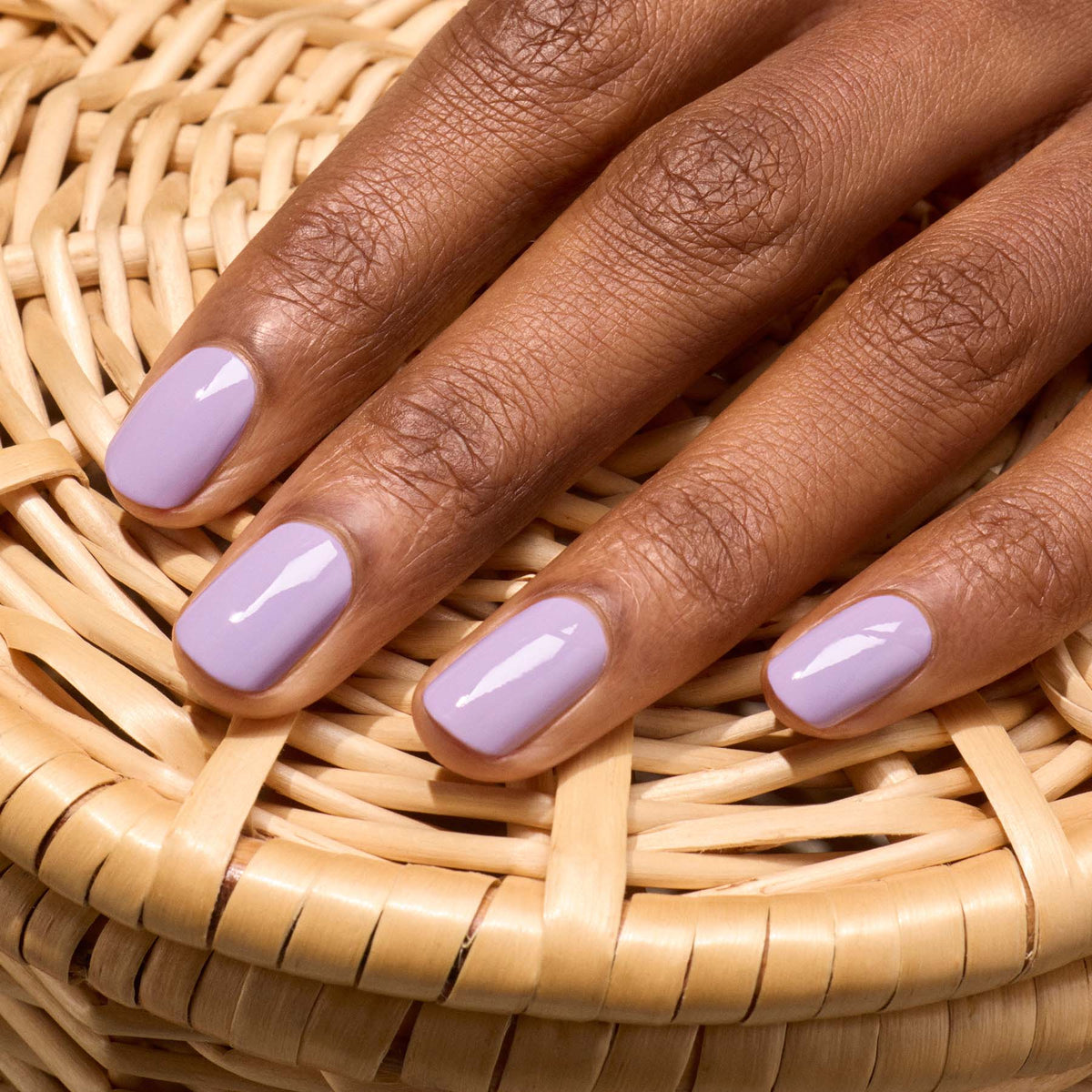 A hand with short, manicured nails painted in Manucurist’s Lisa Lilas glossy polish rests on a wicker surface, giving off subtle 90s vibes.