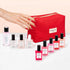 A hand with red nails reaches for a red Manucurist Super Starter Kit - Jelly Nail Polish organic cotton pouch, surrounded by various Manucurist gel-effect nail polishes and a nail polish remover bottle on a white surface.