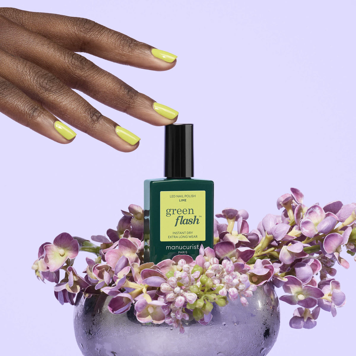 A hand with lime green Manucurist Lime nails reaches for an LED gel polish bottle, surrounded by purple lilacs in a clear bowl on a soft lavender background.