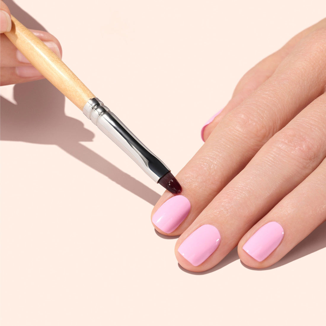 A hand with neatly manicured nails painted light pink is being detailed with the Manucurist Flat Brush, held by another hand, all set against a pale background to highlight meticulous cuticle care and precision in manicure.