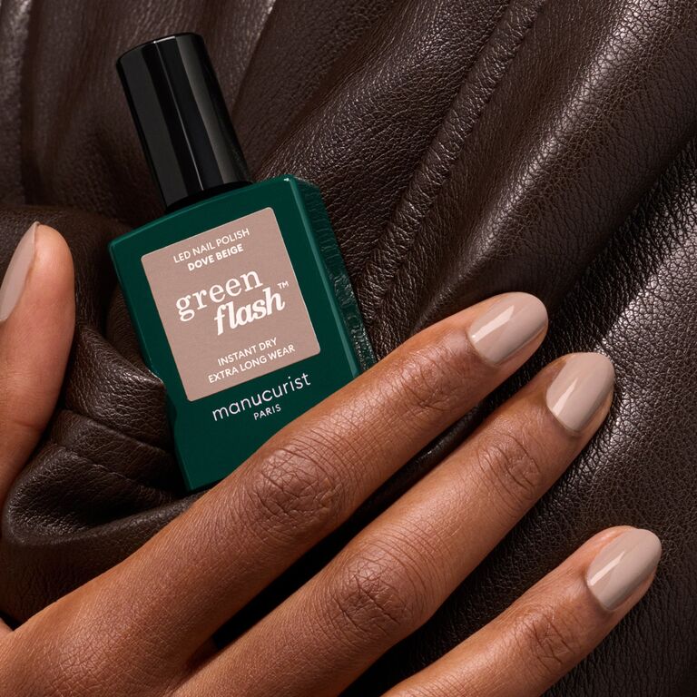 A hand with neatly manicured pastel beige nails holds a bottle of Manucurist Dove Beige nail polish against brown leather.