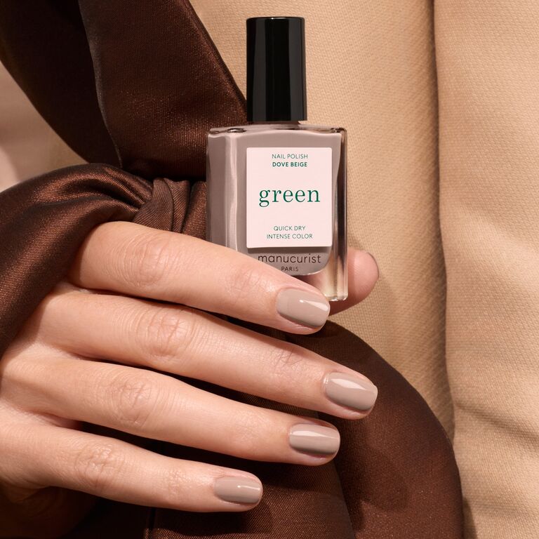 A hand with pastel beige manicured nails holds a bottle of Manucurist Dove Beige nail polish, with a brown satin ribbon draped around the hand.
