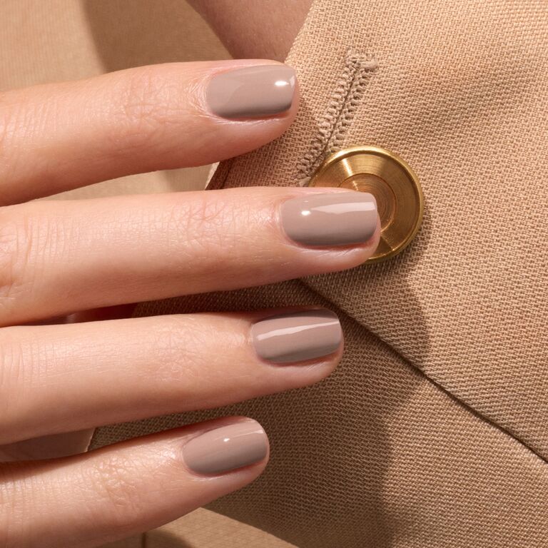 A hand with short, neatly manicured nails painted in Dove Beige by Manucurist rests on a beige fabric sleeve with a gold button.