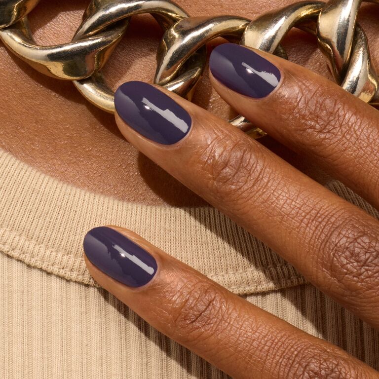 A close-up of a hand with short, oval-shaped nails painted in Manucurist’s Queen of Night, a deep purple polish, rests on beige ribbed fabric with a chunky gold chain necklace in the background.