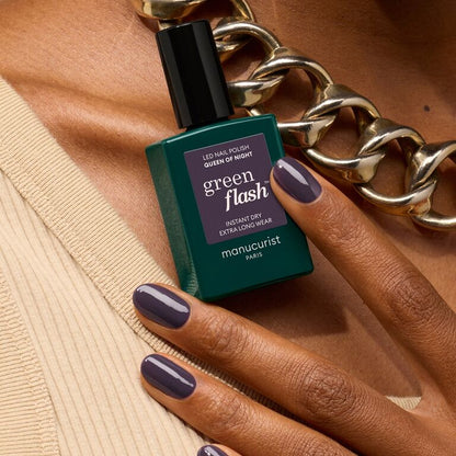 A hand with dark violet gel nail polish, Queen of Night by Manucurist, holds a green bottle. The person wears a chunky gold chain necklace and a beige top.