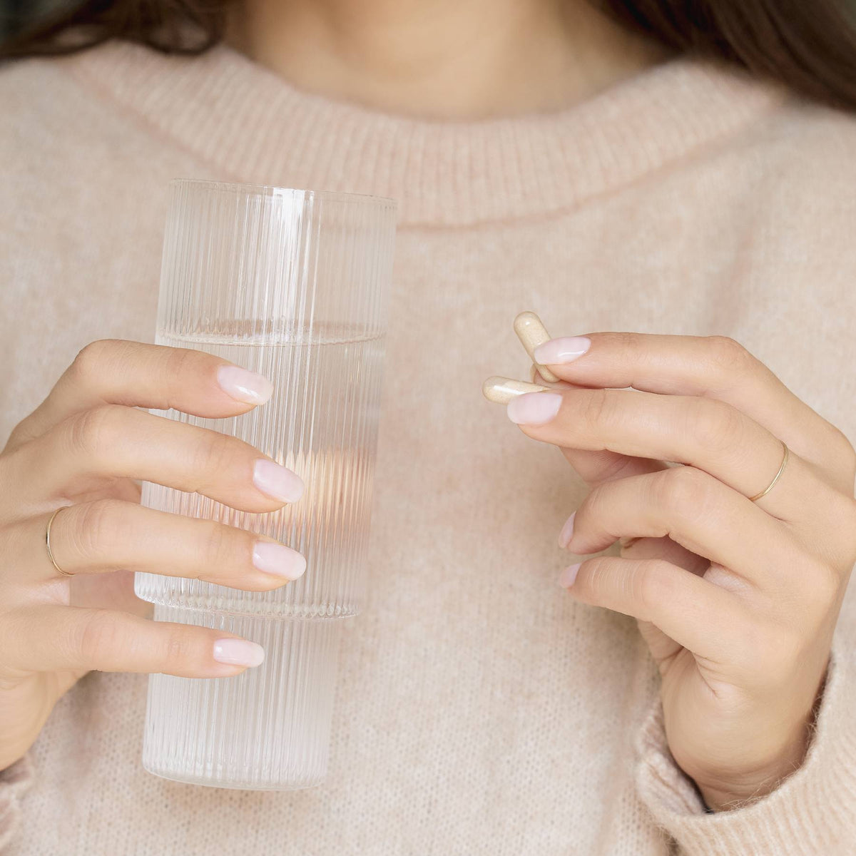 A person in a beige sweater holds a clear, ribbed glass of water and two Manucurist Nail Boost supplements, preparing to take the pills to help strengthen their nails.