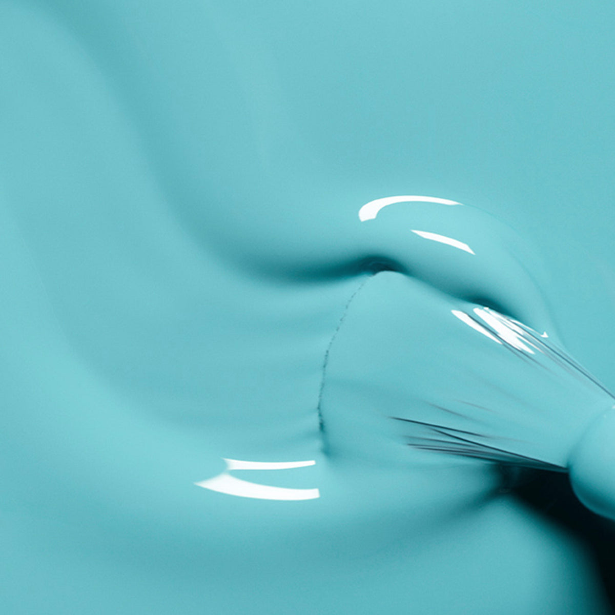 A close-up shows a paintbrush applying Riviera by Manucurist, a smooth, glossy turquoise blue nail polish, with light reflecting off the wet finish to create bright white highlights.