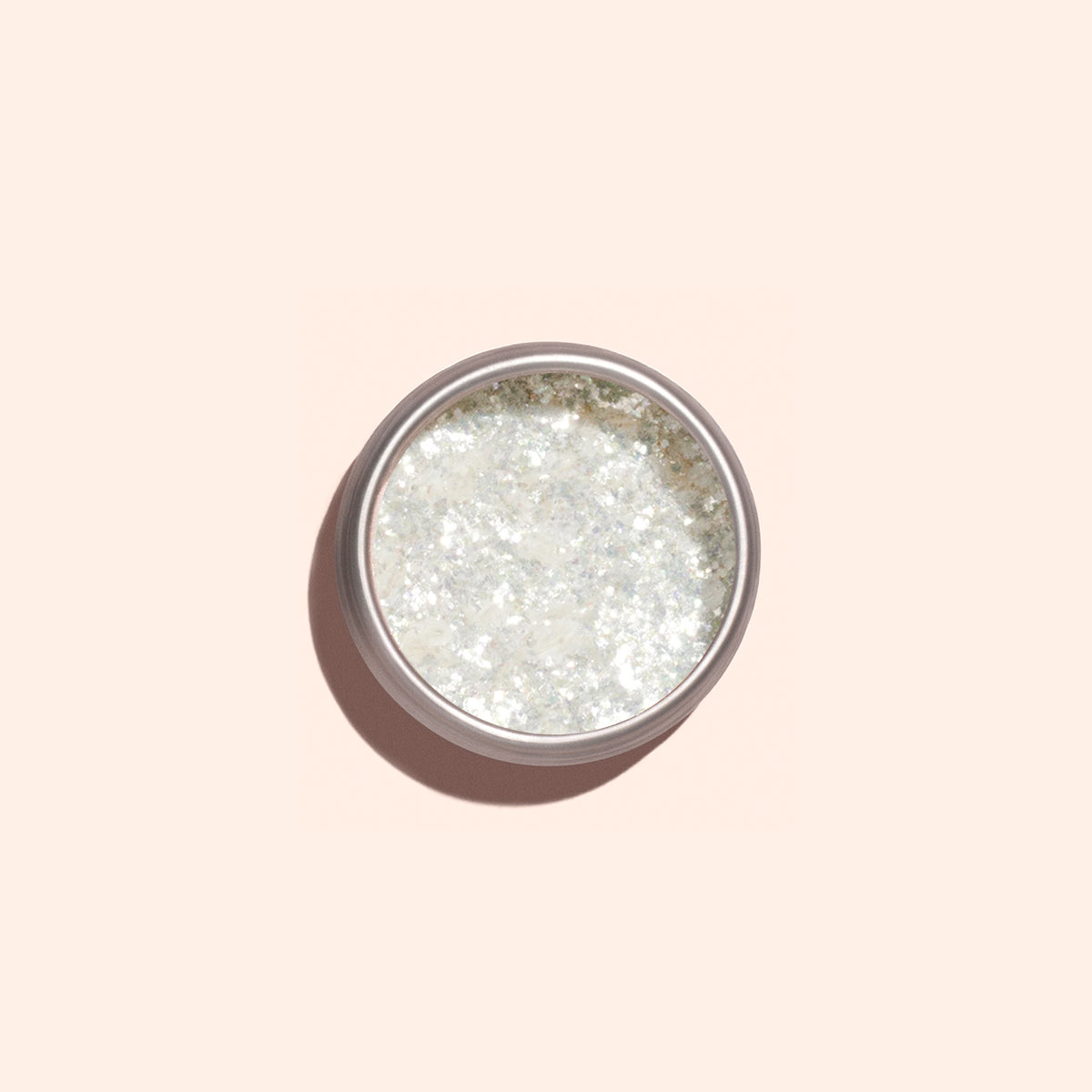 A small round container of Manucurist Nacres Flakes, an iridescent, glittery white cosmetic with pearlescent flakes, sits on a light beige background with a soft shadow.