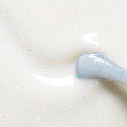 A close-up of a nail polish brush applying Nebula, a milky white polish with tiny colorful glitter specks from Manucurist, on a smooth surface.
