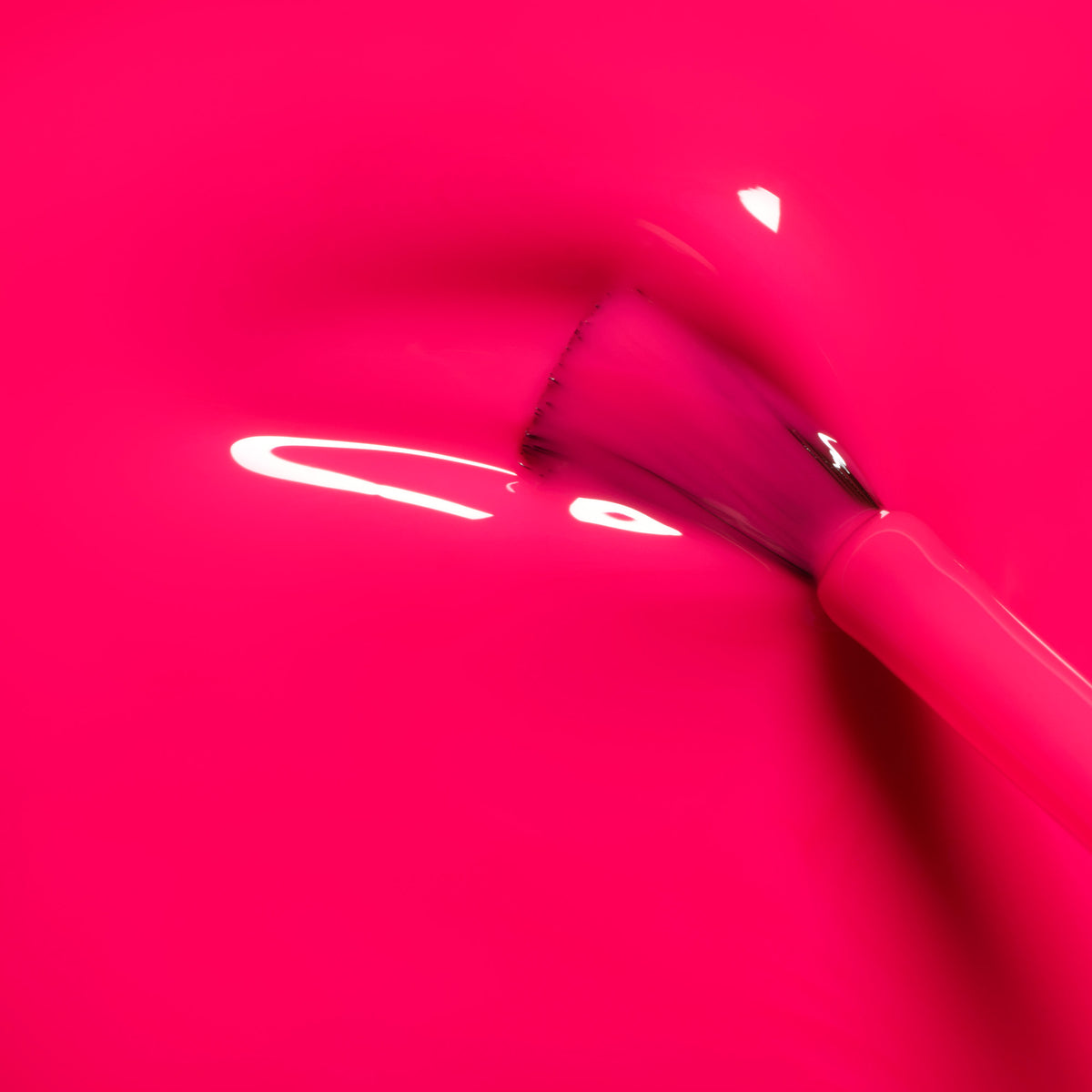 A Manucurist Neon Pink makeup brush glides through glossy neon pink nail polish, creating smooth, light-reflecting strokes for a vibrant summer look.