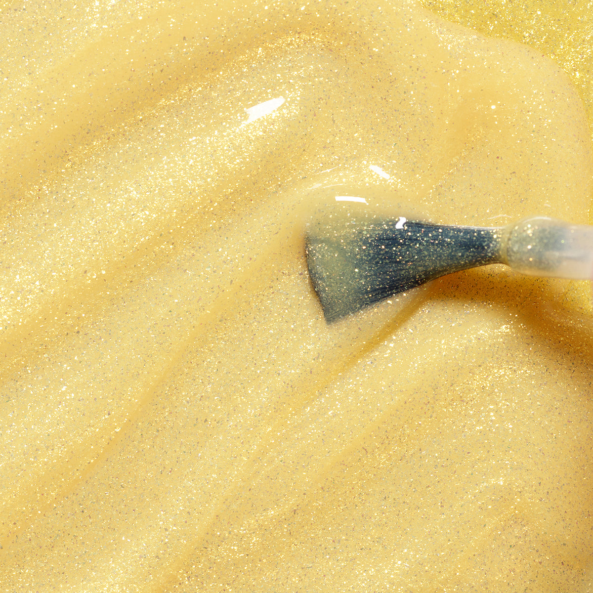 A close-up of a brush applying Pia the Bee, a yellow glitter nail polish by Manucurist made with naturally sourced ingredients, across a smooth surface.