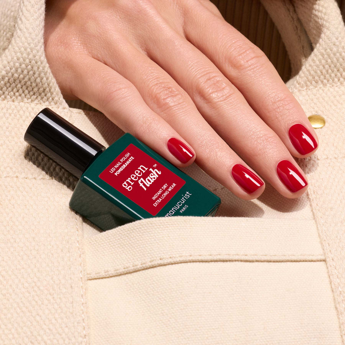 A hand with glossy pomegranate red nails holds a Manucurist Green Flash "Pomegranate" gel polish bottle against beige textured fabric, partially tucked into a pocket.