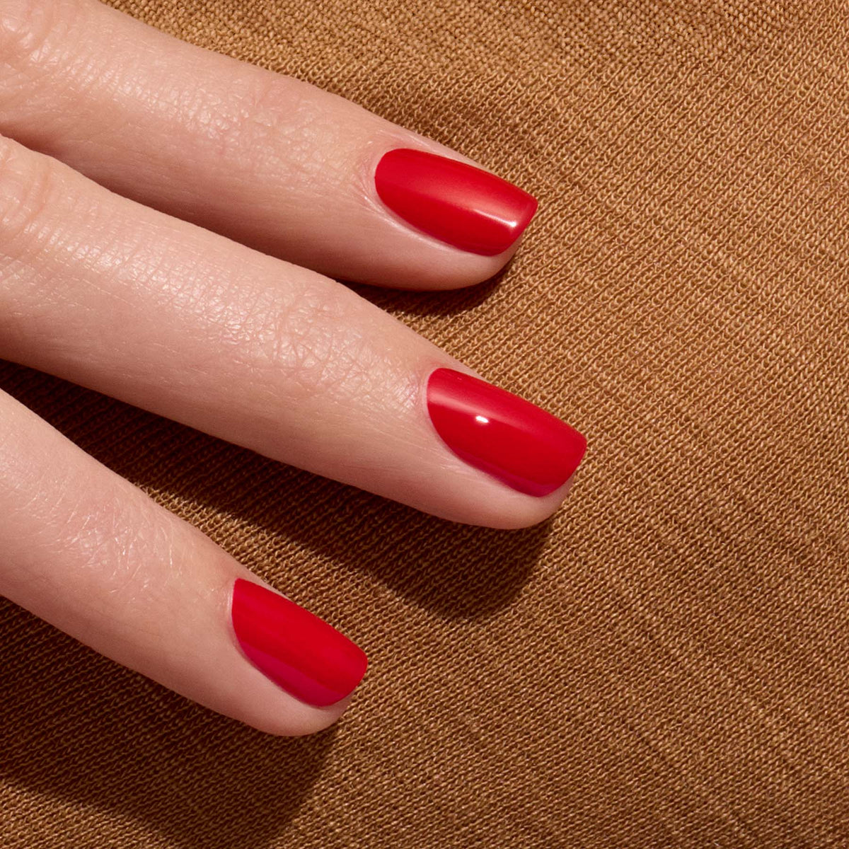 A close-up of a hand with neatly manicured nails painted in Poppy Red - Free by Manucurist-test, resting on a textured brown fabric surface.