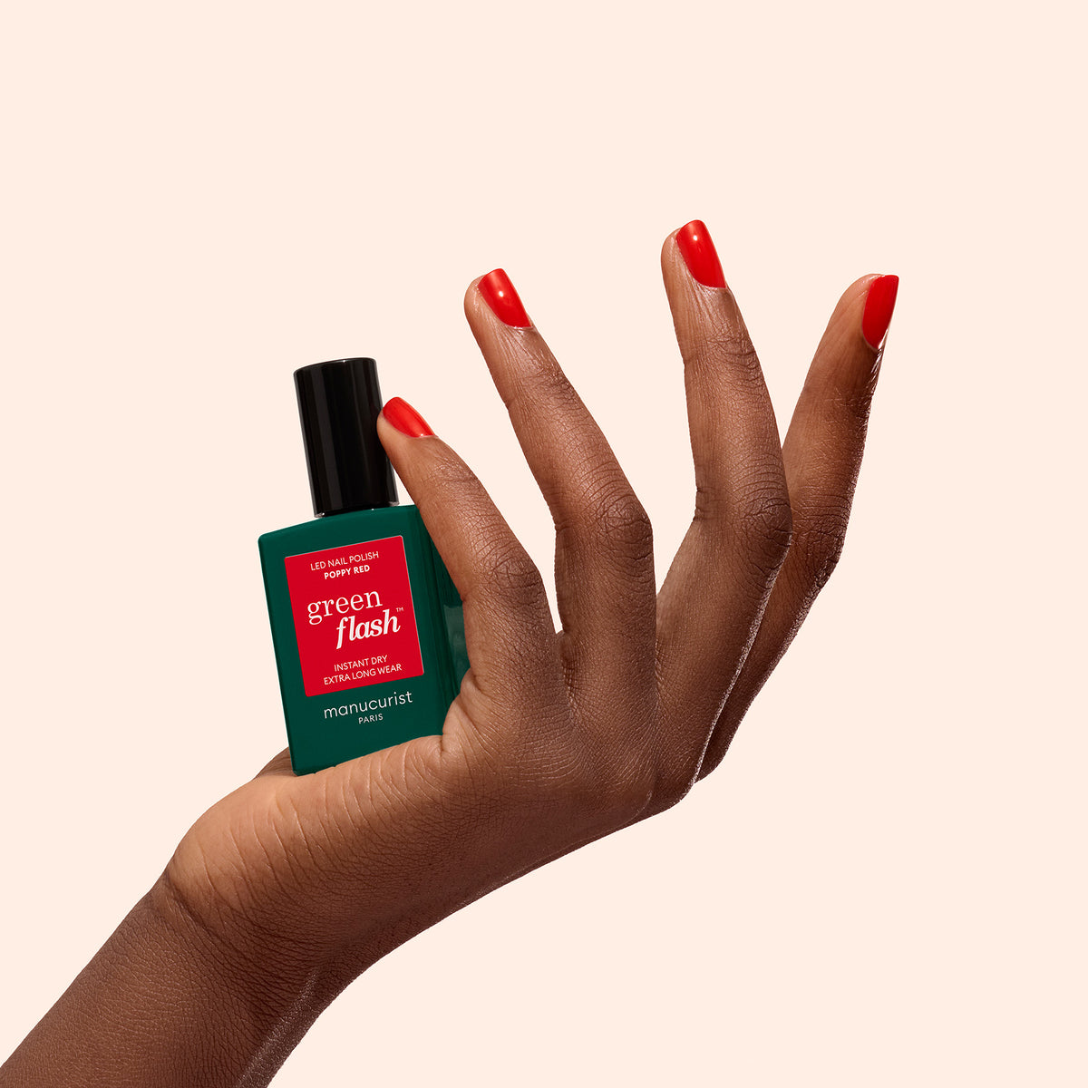 A hand with orangey red nail polish holds a bottle of Manucurist-test Poppy Red - Free gel polish against a light beige background.