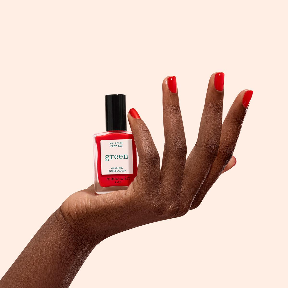 A hand with neatly manicured poppy red nails holds a bottle of Manucurist-test Poppy Red - Free nail polish against a plain light beige background.