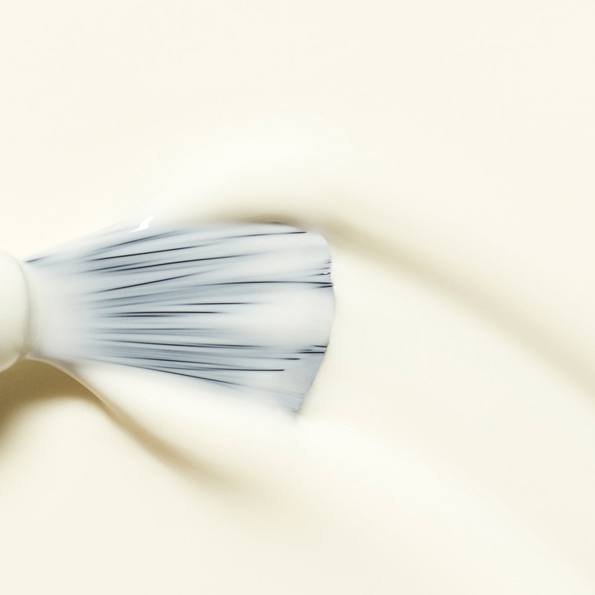 A close-up of a brush with white bristles gliding Manucurist Porcelaine gel polish across a light surface, creating a smooth, glossy finish.