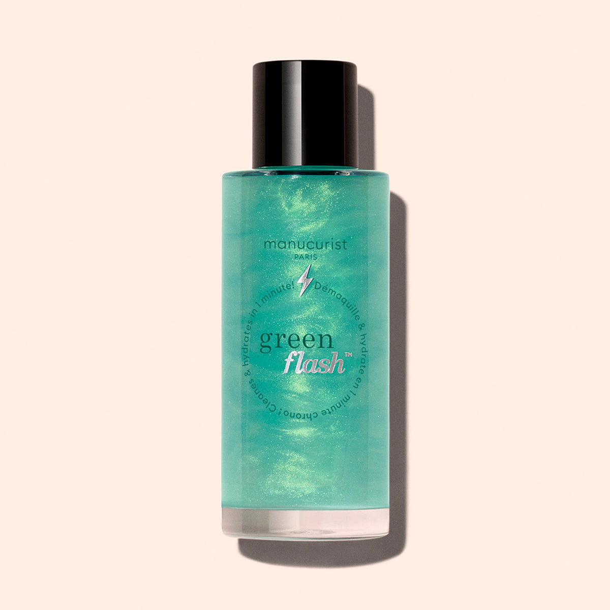 A bottle of Manucurist Green Flash™ Gentle Glitter Remover features a black cap, teal-green liquid, and a minimalist label, photographed against a light beige background.